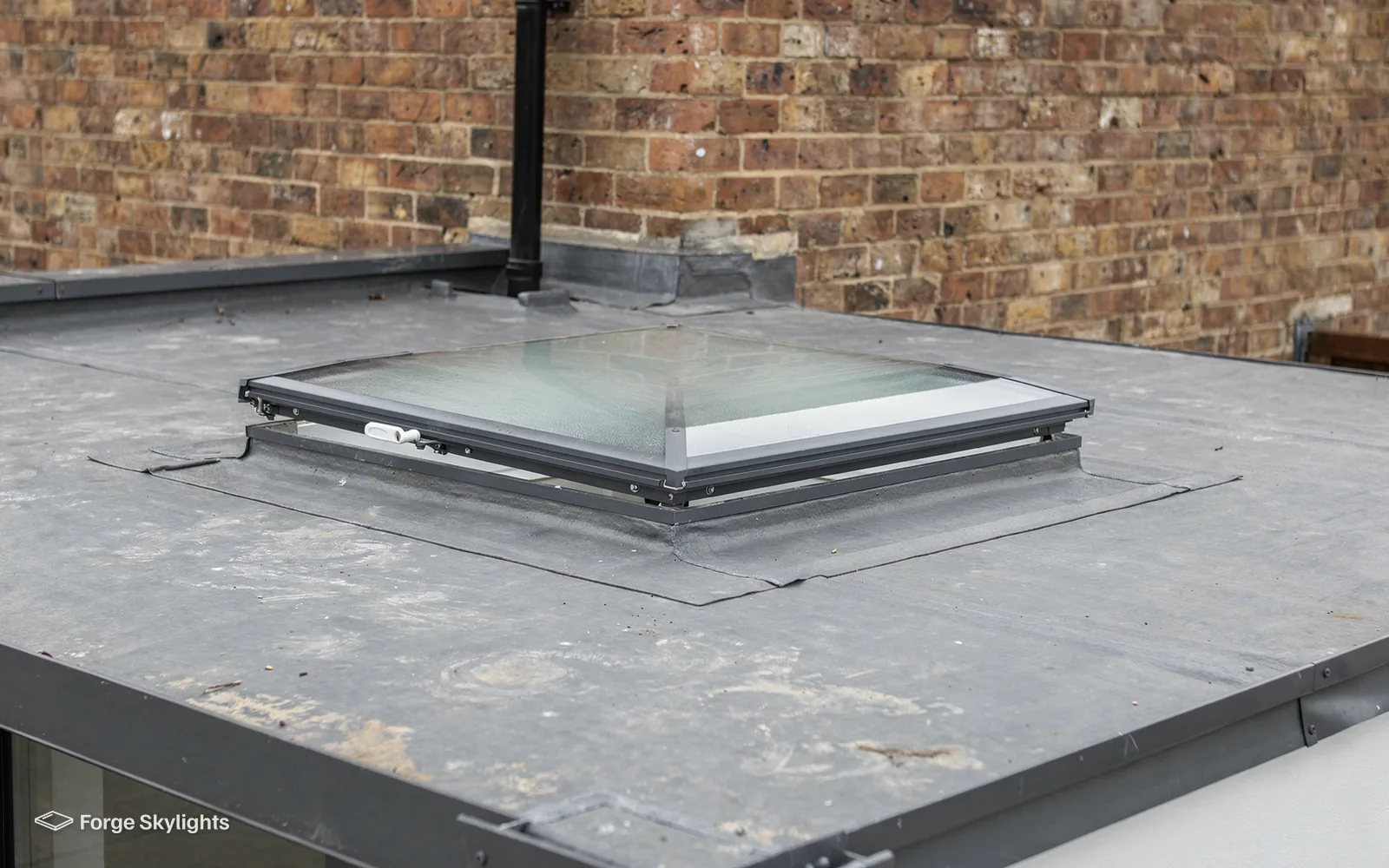 A closed flat rooflight on a felt roof beside a brick wall