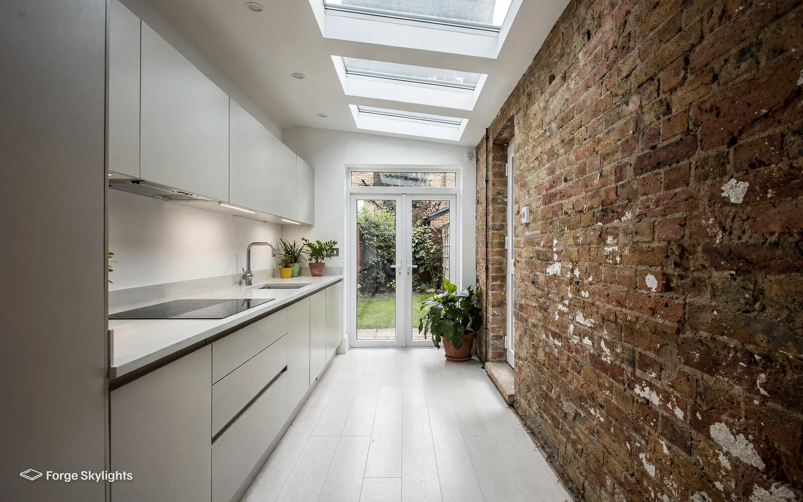 A narrow galley kitchen beside an exposed brick wall, with a row of rooflights along the ceiling