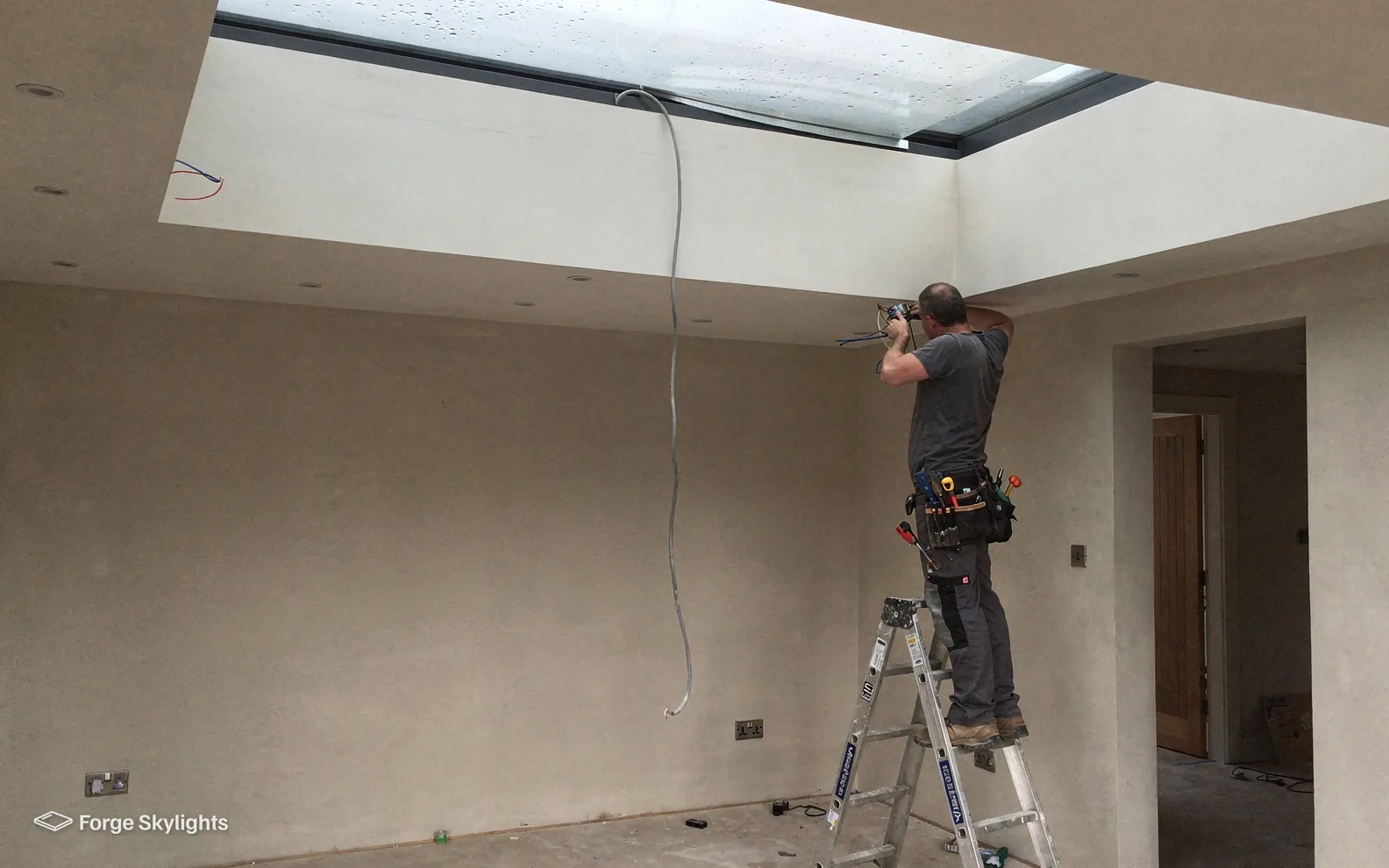 An electrician on a stepladder works under a flat rooflight in a freshly plastered room