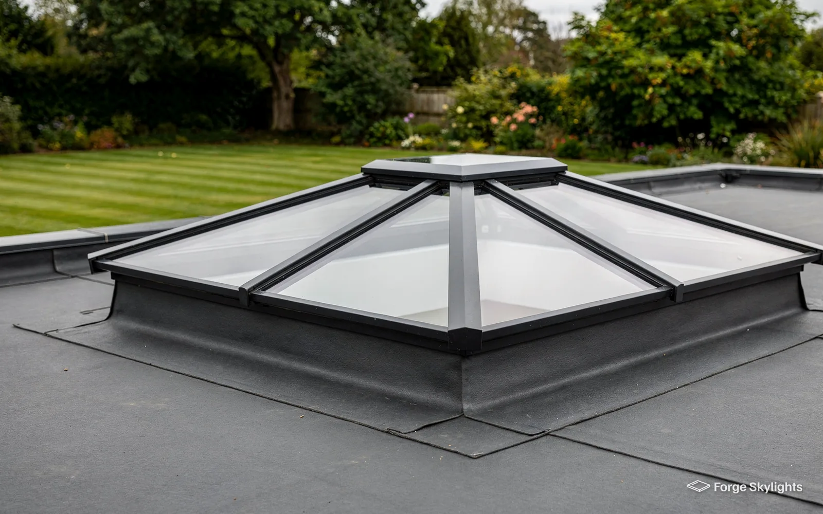 Glass roof lantern with hipped ends rising from a flat roof, trees in the garden beyond