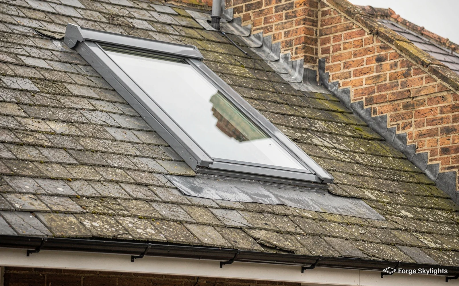 A roof window set in a weathered slate roof beside a brick chimney
