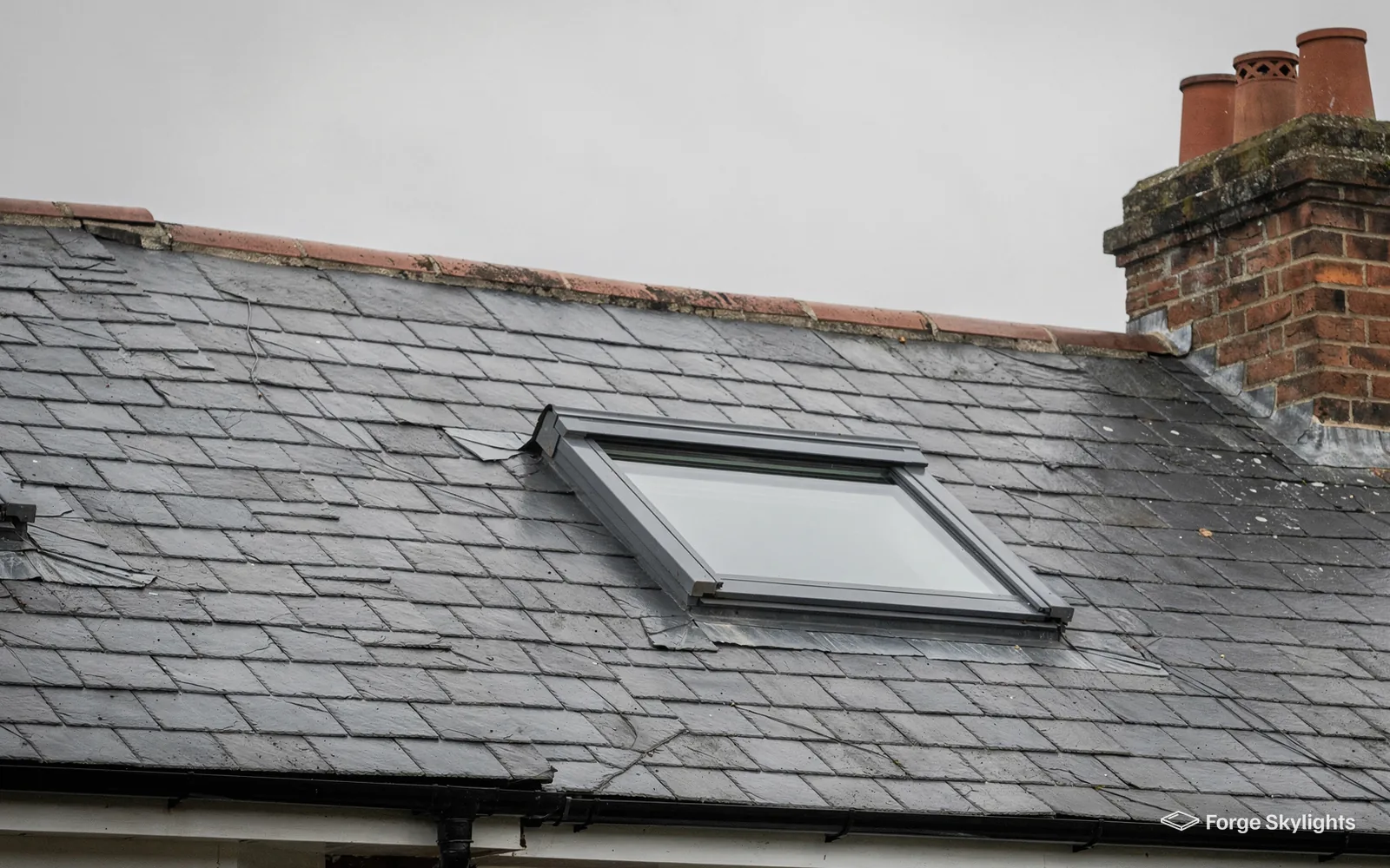 A roof window in a grey slate roof below a chimney with clay pots