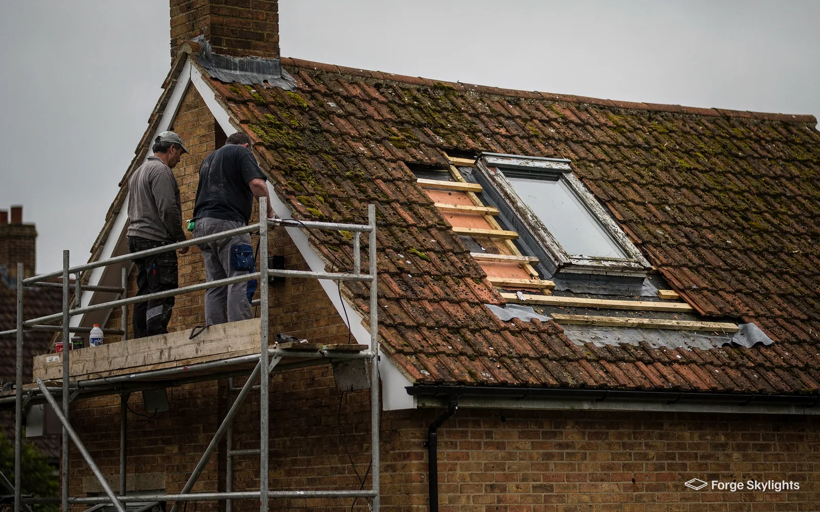 Two fitters on scaffolding fit a roof window into a clay tile roof with the tiles stripped around the opening
